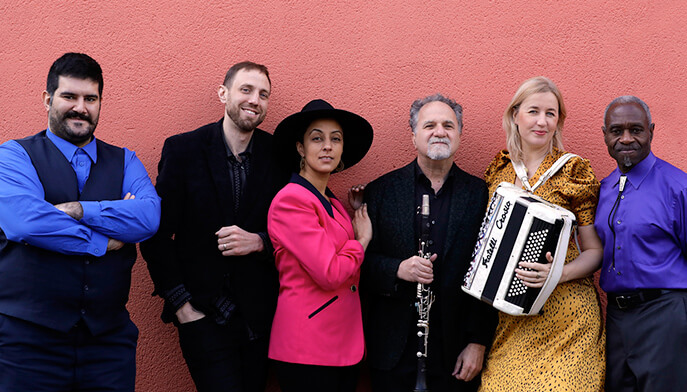 A diverse group of individuals stands together, smiling, with one person playing an accordion among them.