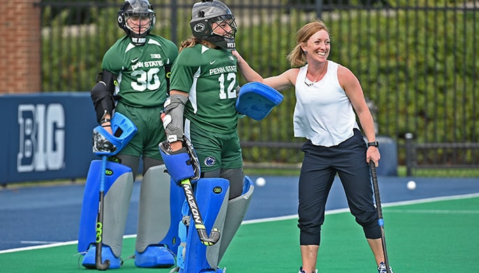 Field Hockey coach smiling as she poses for a candid photo with two of her geared-up student-athletes.