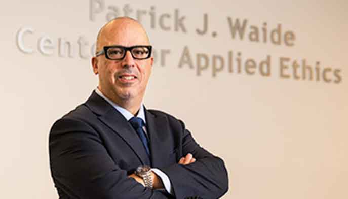 Gregg Caruso stands in front of a wall that says Patrick J. Waide Center for Applied Ethics