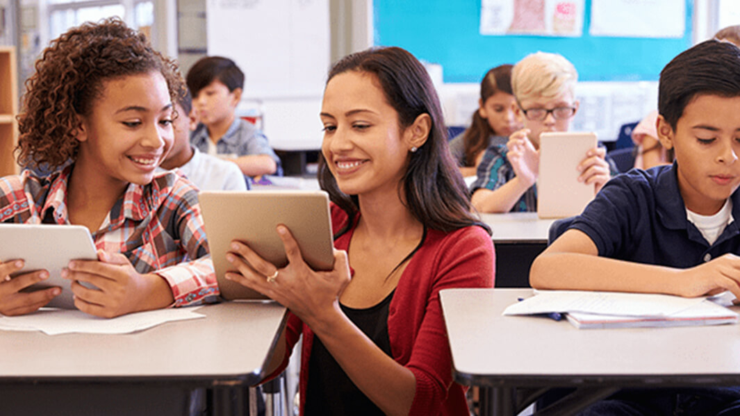 Children and a teacher gather around a tablet, exploring educational content together in a classroom setting. 