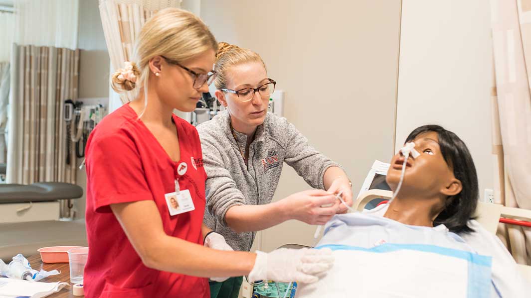 One student in red scrubs and a professor in front of a simulation mannequin.