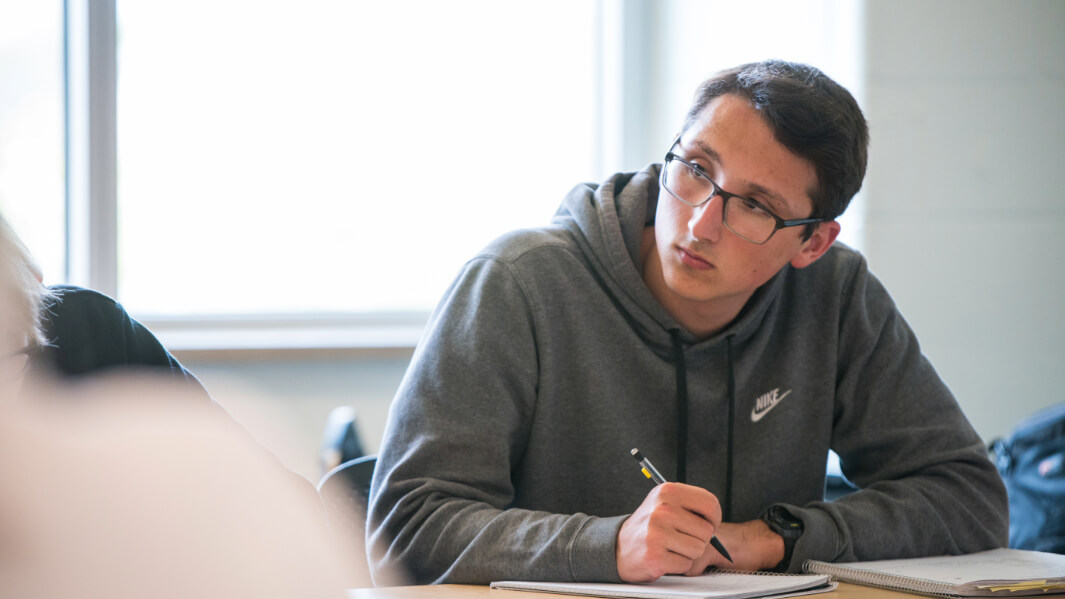 A student writes with a pencil in a notebook at a desk.