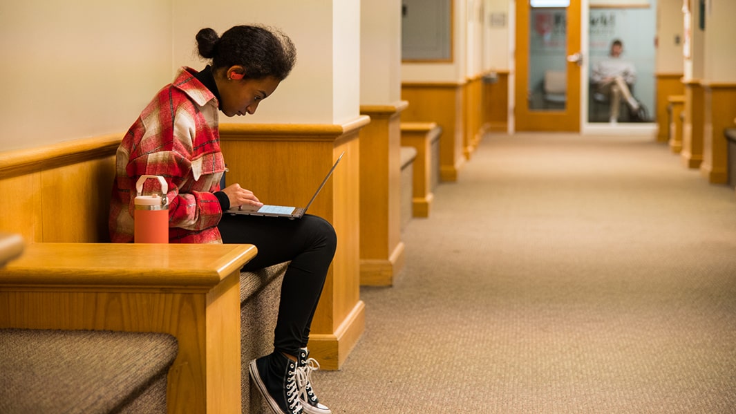 A student sits on a bench indoors using a laptop on their lap.