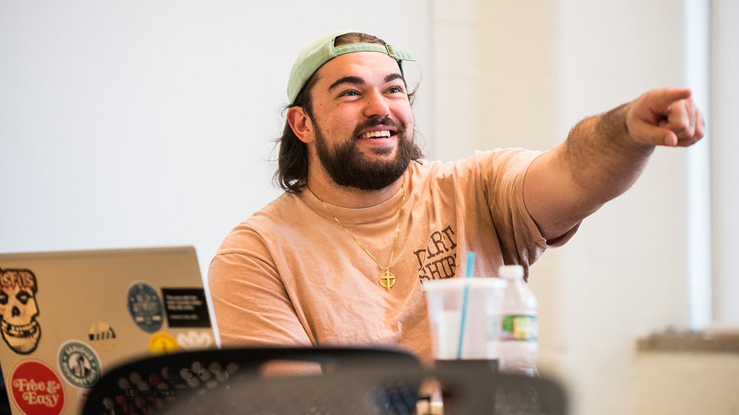 A student smiles and points while sitting at a desk.