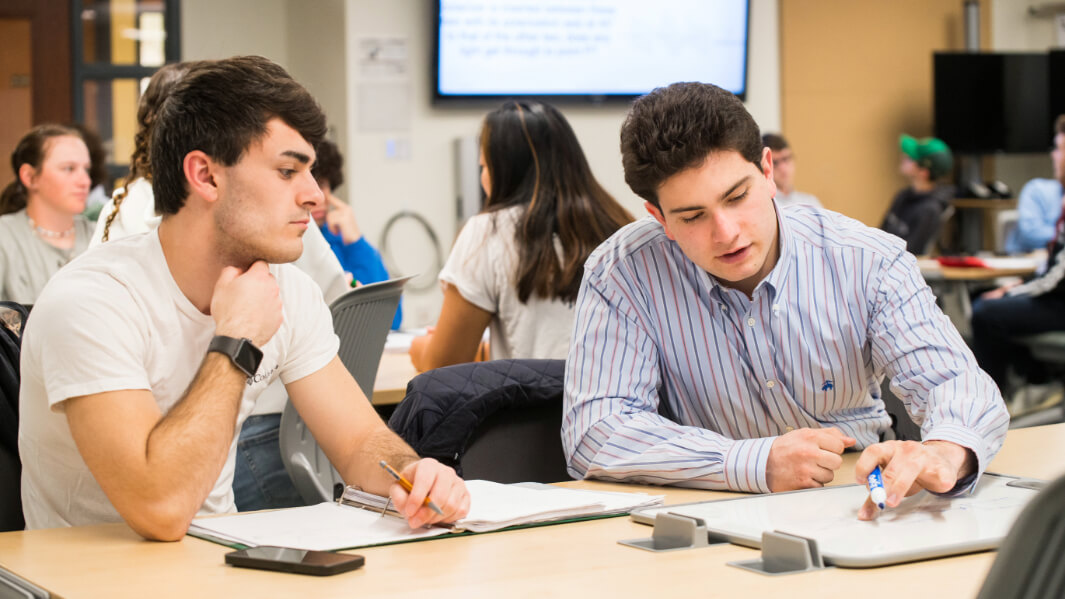 Two students sit at a table solving a problem on a whiteboard.