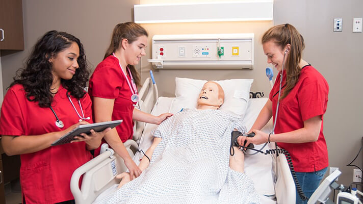 Students in red scrubs stand around a manikin in bed.