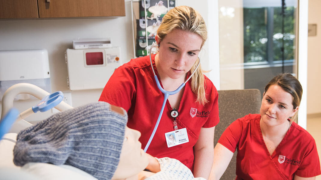 Two students in red scrubs work on a manikin.