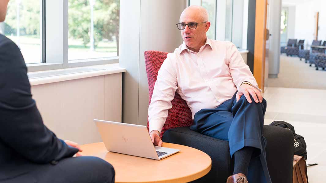 A professor sits in a chair while speaking to a student.