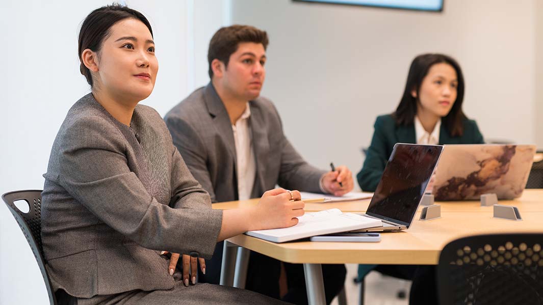 Three students sit at a table covered in notebooks and laptops.