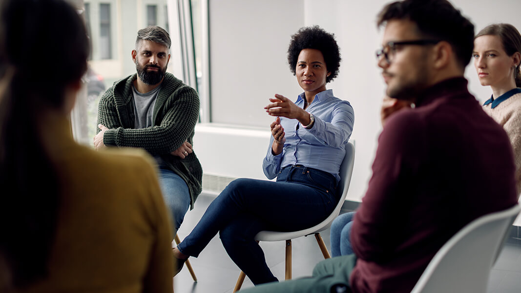 Students sitting in a circle of chairs in a classroom speaking to each other and gesturing.