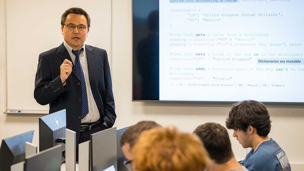 A professor in a suit and tie addresses a group of attentive students in a classroom setting.