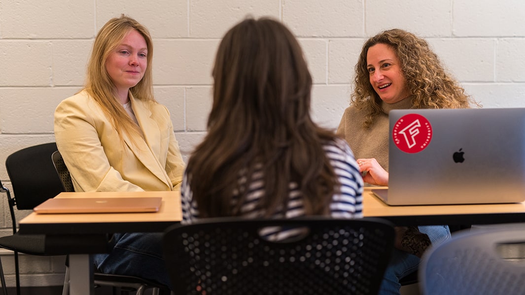 Two students sit at a table facing another student.