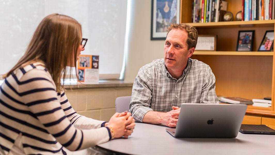 A student and professor sit in front of an open laptop and engage in conversation.