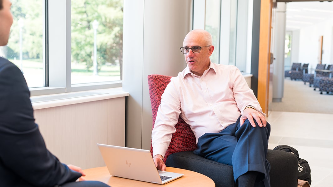 A professor sits in a chair while speaking to a student.