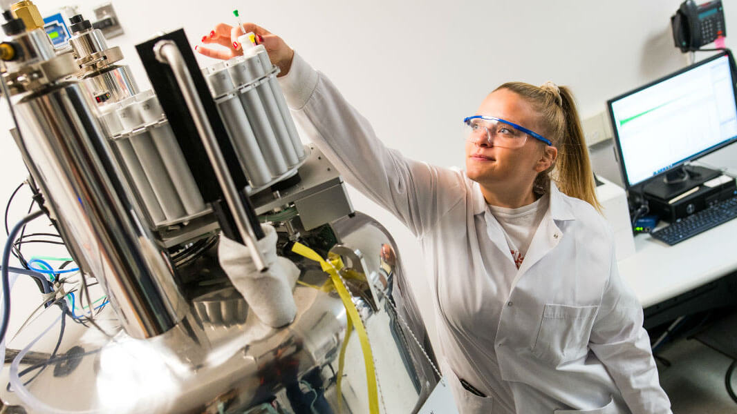 A student wearing a labcoat and eye protection is reaching up to a complex looking machine to take out a test tube.