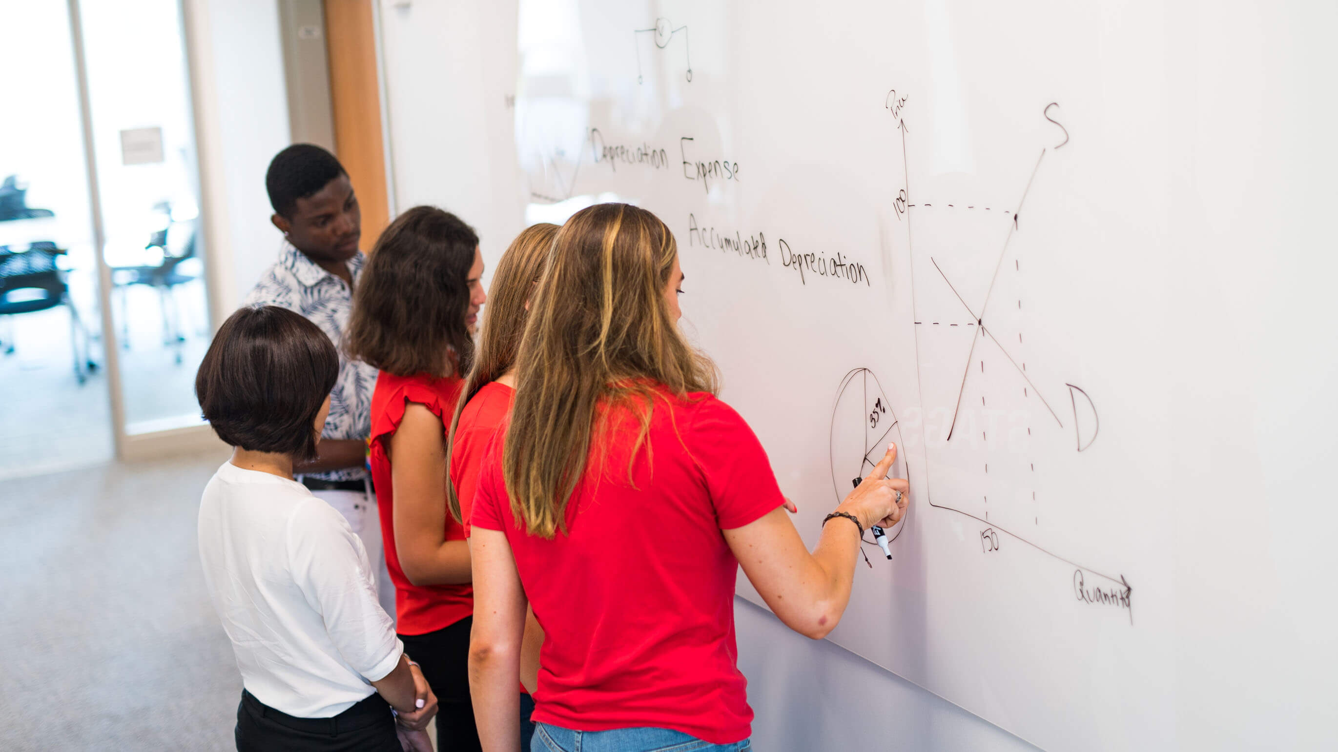 4 business students work on a problem together on a white board with a professor supervises.