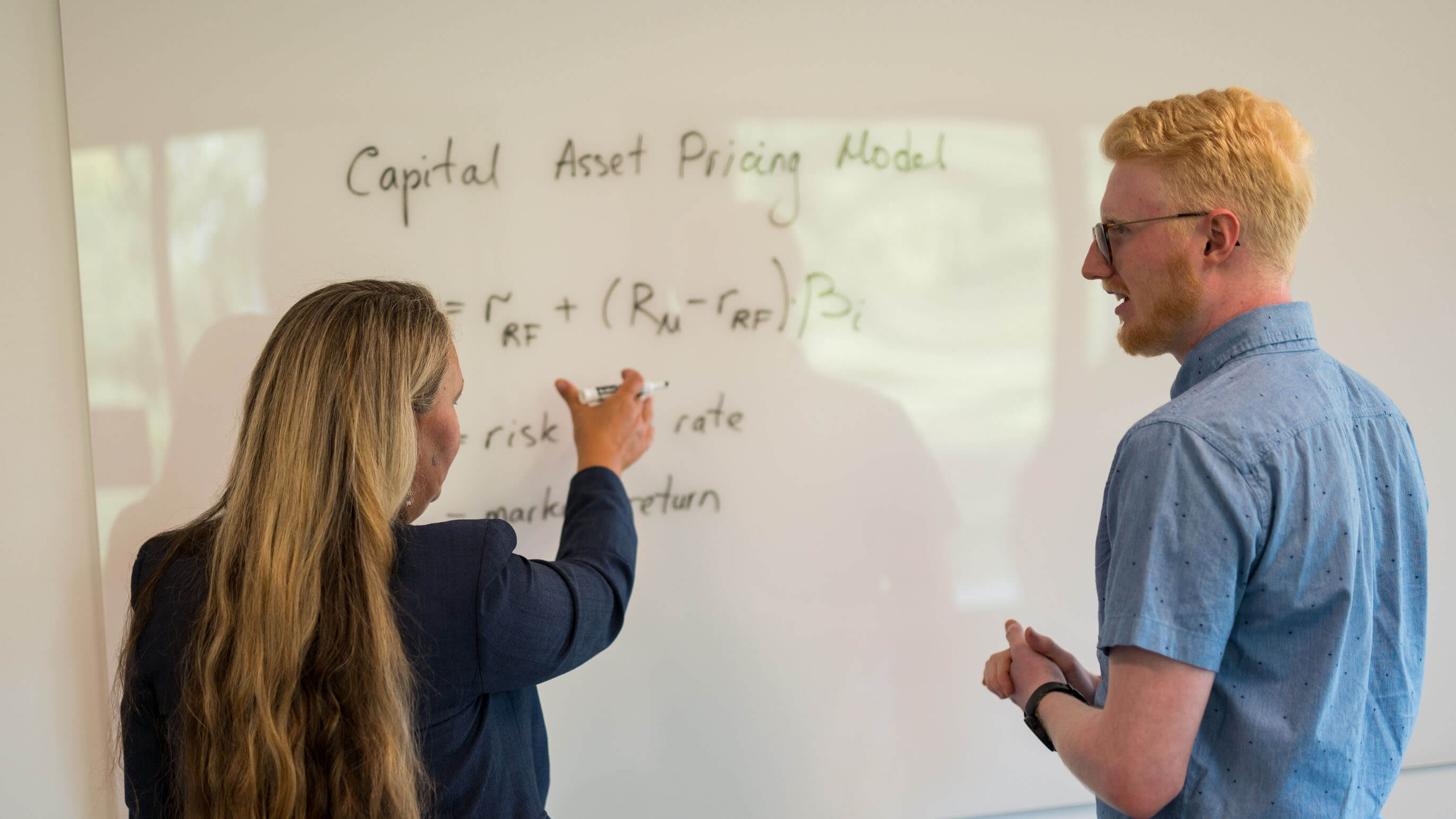 A professor walks a student through a problem on a whiteboard.