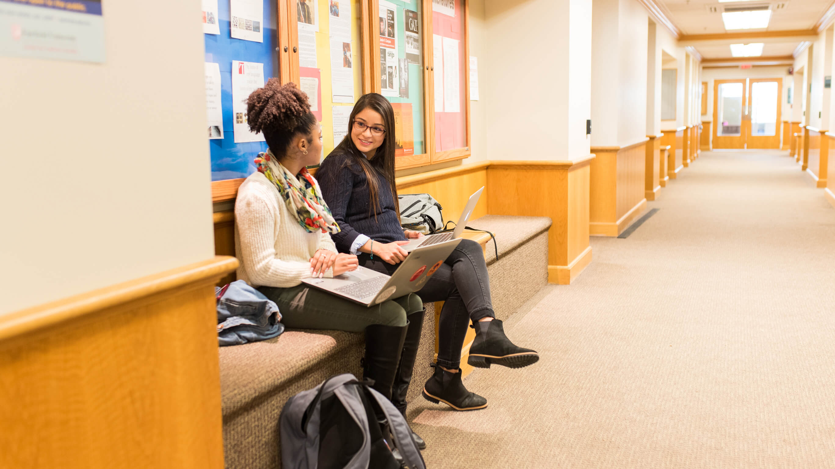 Two students sit on a bench indoors.