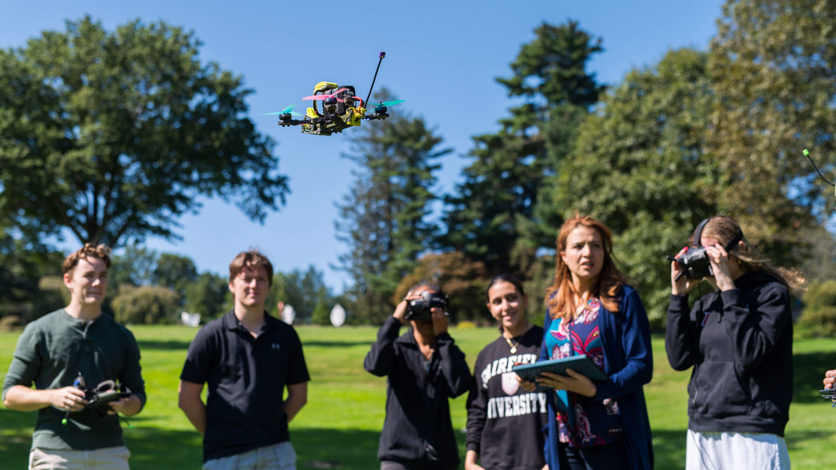 Students standing in a circle with their professor who is operating a drone in the middle of all of them. They are standing on bright green grass with trees surrounding them and a blue sky in the background. 