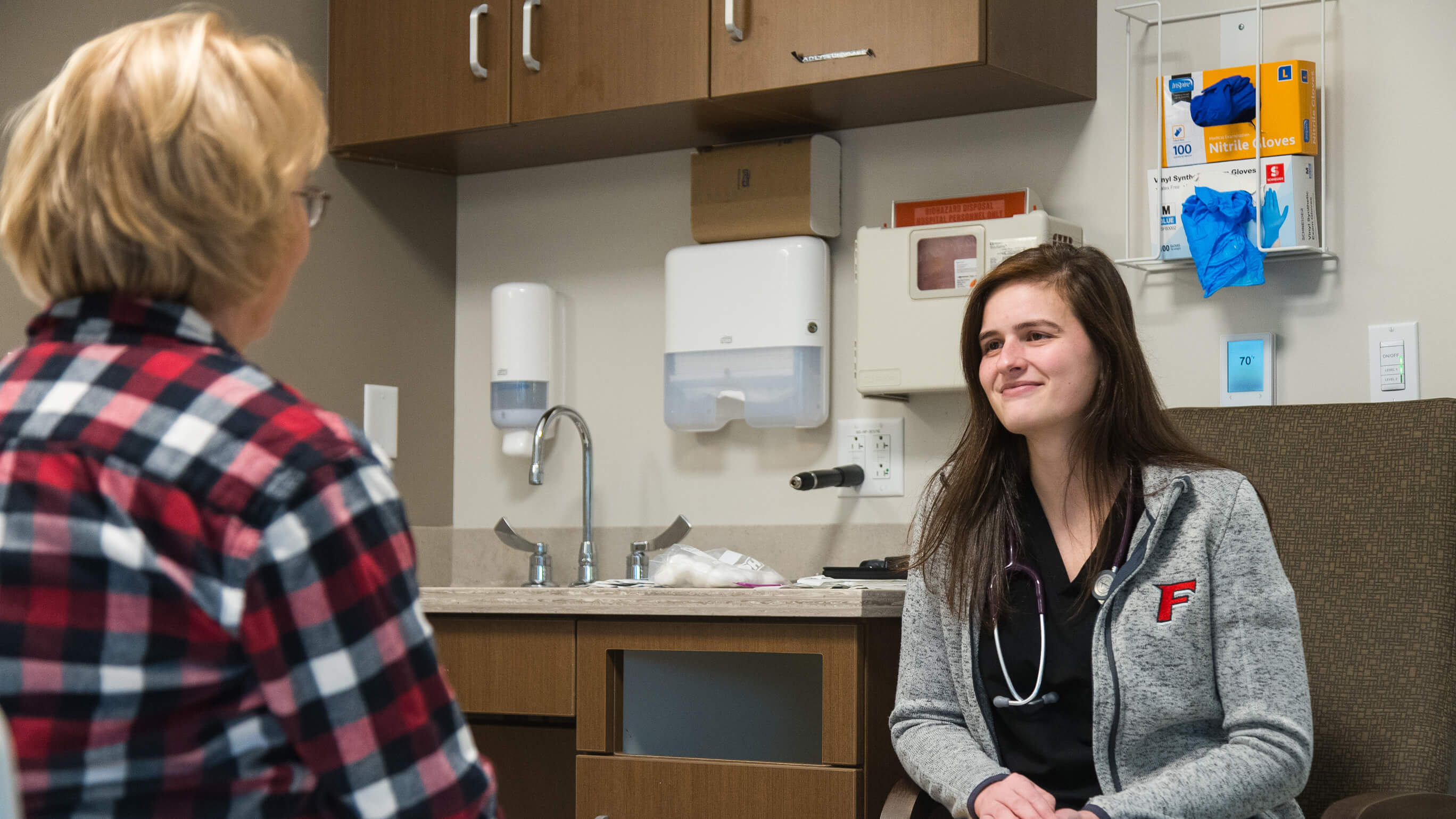 A student in a Fairfield jacket with a stethoscope around her neck talks to a patient.