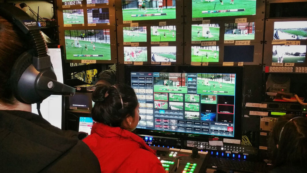 Two students in headsets sit in front of an editing bay displaying sports footage.