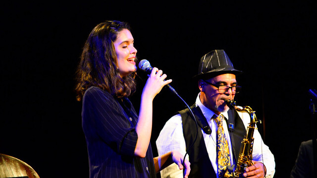 A person sings into a microphone in front of a saxophone player.