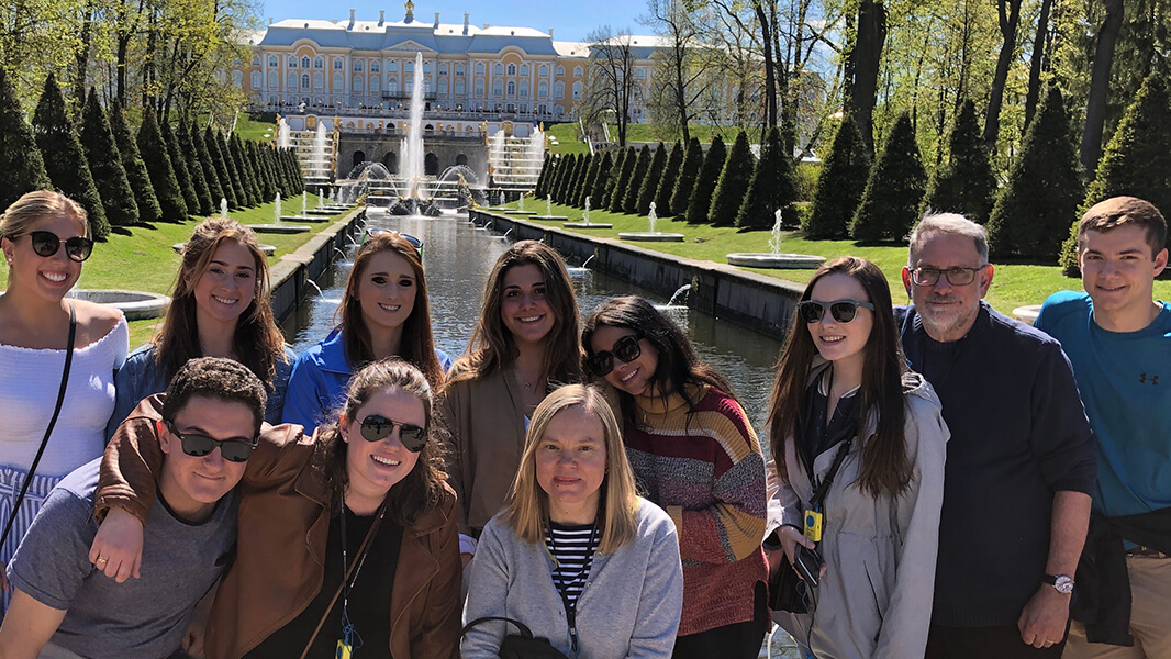 A group of students smiles outdoors in front of a large fountain pool and historical building. 