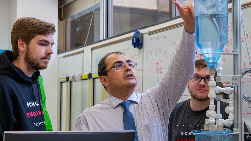 Engineering professor standing between two students pointing something out on a machine that holds a tank of clear liquid. 