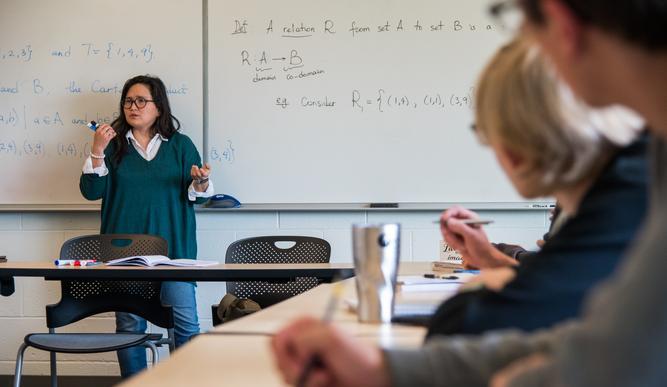 A professor speaks in front of a white board in a small classroom