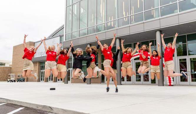 A group of students in red shirts are pictured mid-jump in front of a glass building.