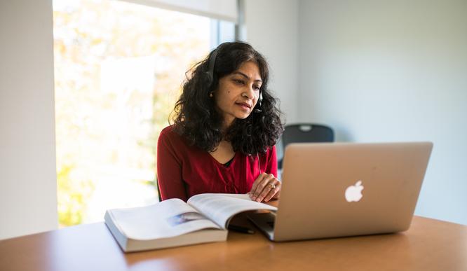 A woman dark hair and headphones works at a laptop with a book open in front of her