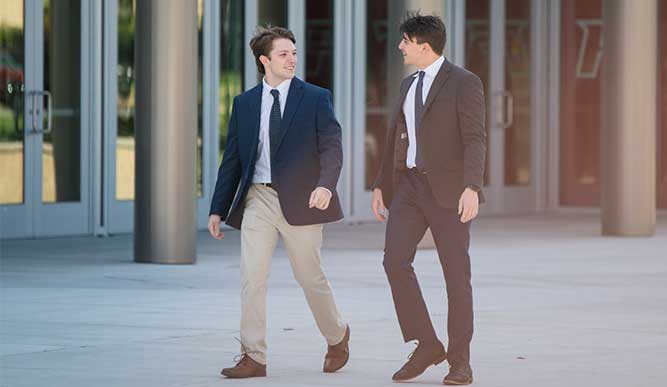 Two students wearing suits walking in front of a building on campus talking and smiling at each other. 