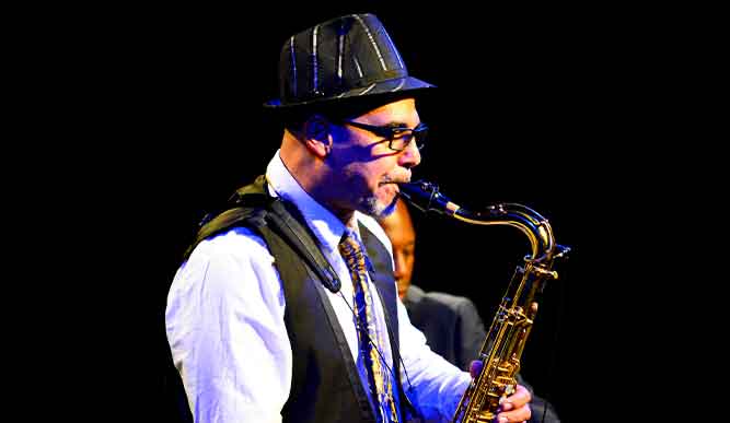 A man wearing a hat and glasses passionately plays a saxophone, showcasing his musical talent and style.