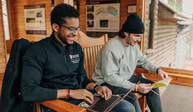 Students sitting on a bench. One is on their computer and another is reading. 