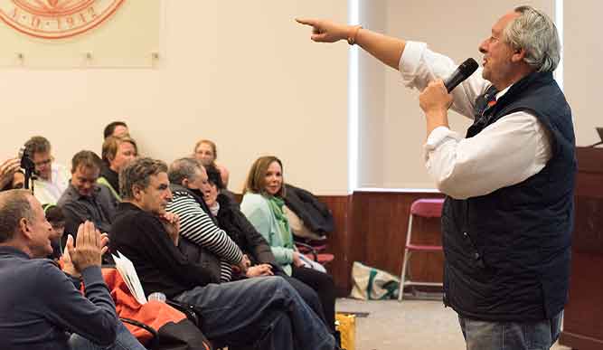 A man addresses an audience in a well-lit room, engaging them with his speech and gestures.