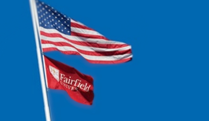 An American flag flies over a Fairfield University flag in front of a blue background.