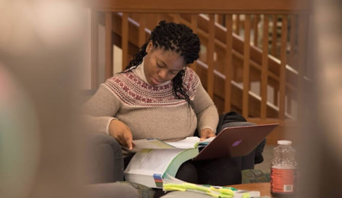 A student reads a textbook while holding a laptop in a library chair.