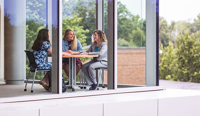 Three women sit at a table by a large window, enjoying a conversation.