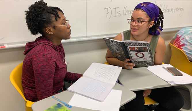 Two students engaged in study at desks, surrounded by books, with a whiteboard in the background.