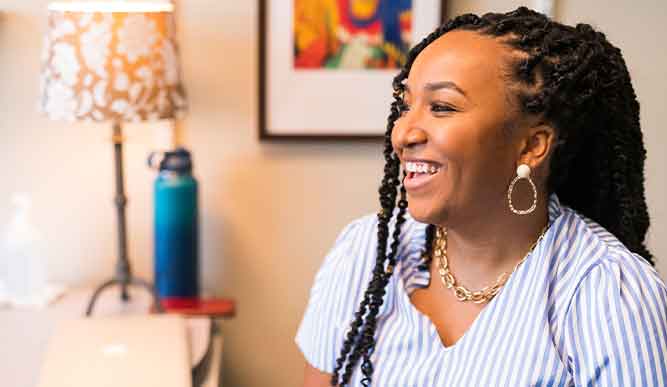 A woman with braids sits at a desk, smiling warmly while engaging in her work.