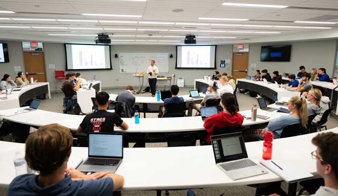 A professors stands in front of a large rounded classroom full of students teaching a class.