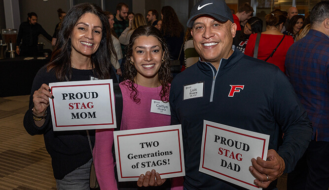 Three people hold up signs: “Proud Stag Mom”, “Proud Stag Dad”, and “Two Generations of Stags!”.