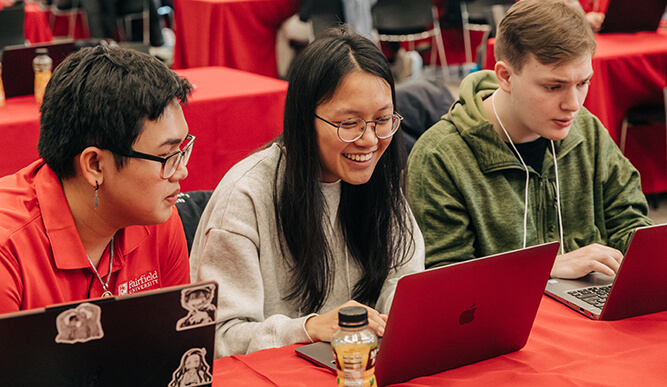 Three students sit in front of open laptops at a red table.