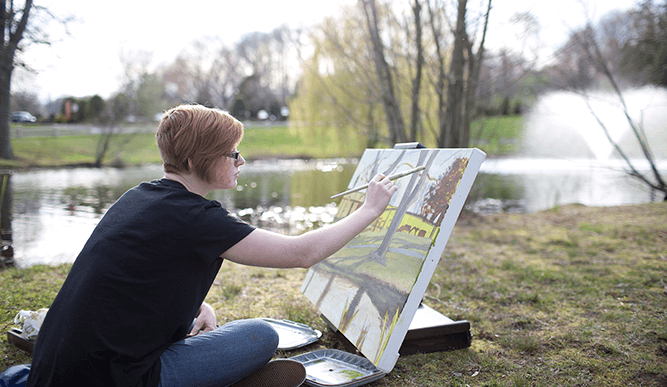 Student paints a still life while sitting on grass around a pond.