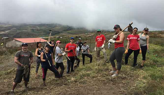 A group of students stand on a hillside holding gardening tools.