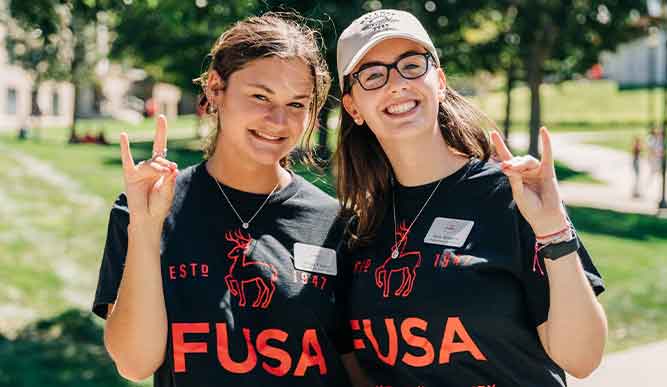 Two young women in fusa shirts smiling and holding up two fingers in a cheerful gesture.