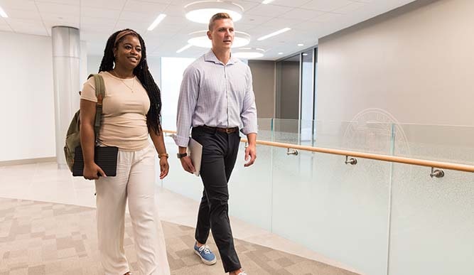 Two students walk down a white hallway holding their laptops.