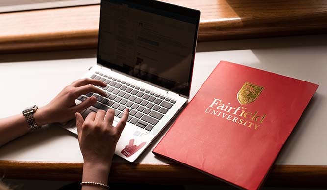 A student uses a laptop which sits next  to a red folder titled “Fairfield University” in gold lettering.