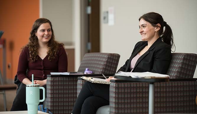 Two students sit in chairs holding notebooks and pencils.