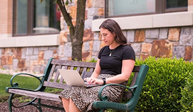 A student sits on an outdoor bench using her laptop with a black tote bag by her feet.